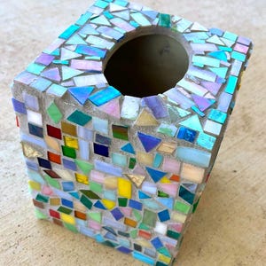 May include: A square tissue box cover featuring a mosaic design made of small, multi-colored glass tiles in shades of blue, green, yellow, pink, and red, set into a light gray grout base with a circular opening on the top surface.