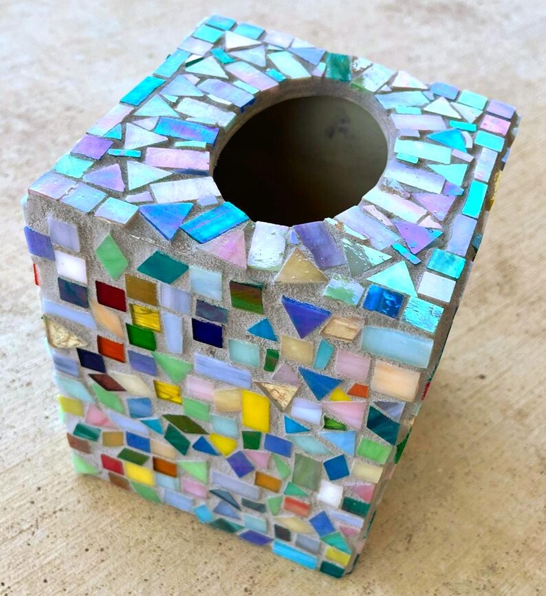 May include: A square tissue box cover featuring a mosaic design made of small, multi-colored glass tiles in shades of blue, green, yellow, pink, and red, set into a light gray grout base with a circular opening on the top surface.