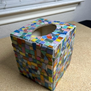 May include: A square tissue box cover decorated with a multi-colored mosaic pattern. The exterior is covered in small, irregularly shaped glass tiles in shades of blue, green, yellow, orange, and pink, set into a light-colored grout base.