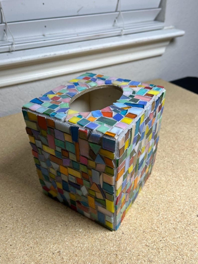 May include: A square tissue box cover decorated with a multi-colored mosaic pattern. The exterior is covered in small, irregularly shaped glass tiles in shades of blue, green, yellow, orange, and pink, set into a light-colored grout base.