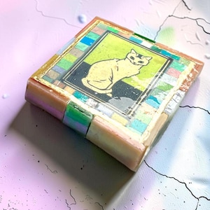 May include: A square decorative tile featuring a central illustration of a seated cat in yellow against a black background. The image is framed by a border of small, multicolored square tiles in shades of blue, green, pink, and gold leaf accents.
