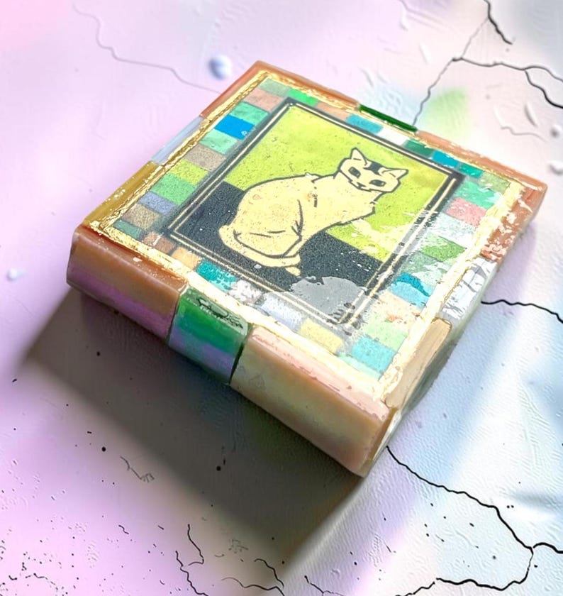 May include: A square decorative tile featuring a central illustration of a seated cat in yellow against a black background. The image is framed by a border of small, multicolored square tiles in shades of blue, green, pink, and gold leaf accents.