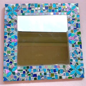 Puede incluir: Un espejo de pared cuadrado con un marco de mosaico decorativo compuesto por peque&ntilde;as piezas de vidrio de formas irregulares en tonos azules, verdes y rosas. Las cuatro esquinas del marco presentan motivos florales de cuatro p&eacute;talos.