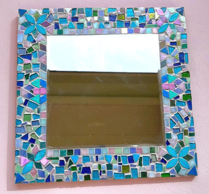 Puede incluir: Un espejo de pared cuadrado con un marco de mosaico decorativo compuesto por peque&ntilde;as piezas de vidrio de formas irregulares en tonos azules, verdes y rosas. Las cuatro esquinas del marco presentan motivos florales de cuatro p&eacute;talos.