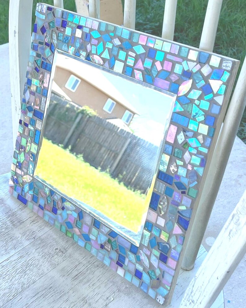 Op de afbeelding: Een vierkante spiegel met een decoratieve lijst bestaande uit talloze kleine, onregelmatig gevormde moza&iuml;ektegeltjes in tinten blauw, lichtroze en iriserend wit, gerangschikt in een dicht patroon rondom het centrale spiegeloppervlak.