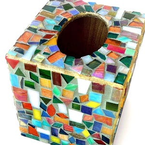 May include: A square tissue box cover featuring a mosaic design made of multicoloured glass shards in shades of red, blue, green, yellow, and white, set in grey grout with a gold-painted top edge and a central oval opening for dispensing tissues.