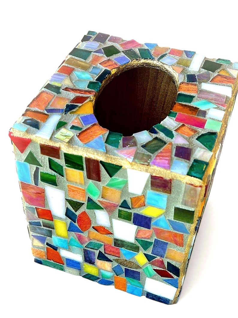 May include: A square tissue box cover featuring a mosaic design made of multicoloured glass shards in shades of red, blue, green, yellow, and white, set in grey grout with a gold-painted top edge and a central oval opening for dispensing tissues.