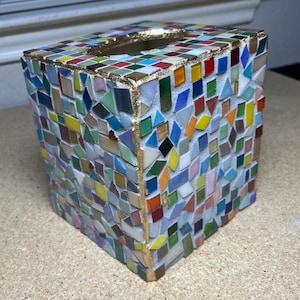 K&ouml;nnte beinhalten: Ein quadratischer Taschentuchbox-&Uuml;berzug, verziert mit einem bunten Mosaik aus kleinen Glasfliesen in Rot, Blau, Gr&uuml;n, Gelb und Wei&szlig;. Die Kanten und die obere &Ouml;ffnung sind mit einem metallisch goldenen Rand versehen, der einen Akzent setzt.