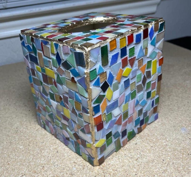 K&ouml;nnte beinhalten: Ein quadratischer Taschentuchbox-&Uuml;berzug, verziert mit einem bunten Mosaik aus kleinen Glasfliesen in Rot, Blau, Gr&uuml;n, Gelb und Wei&szlig;. Die Kanten und die obere &Ouml;ffnung sind mit einem metallisch goldenen Rand versehen, der einen Akzent setzt.