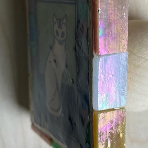 May include: A square mosaic tile featuring a central illustration of a cat on a dark background. The side edge is decorated with three vertically stacked, iridescent, rectangular glass tiles in shades of pink, white, and yellow-gold.