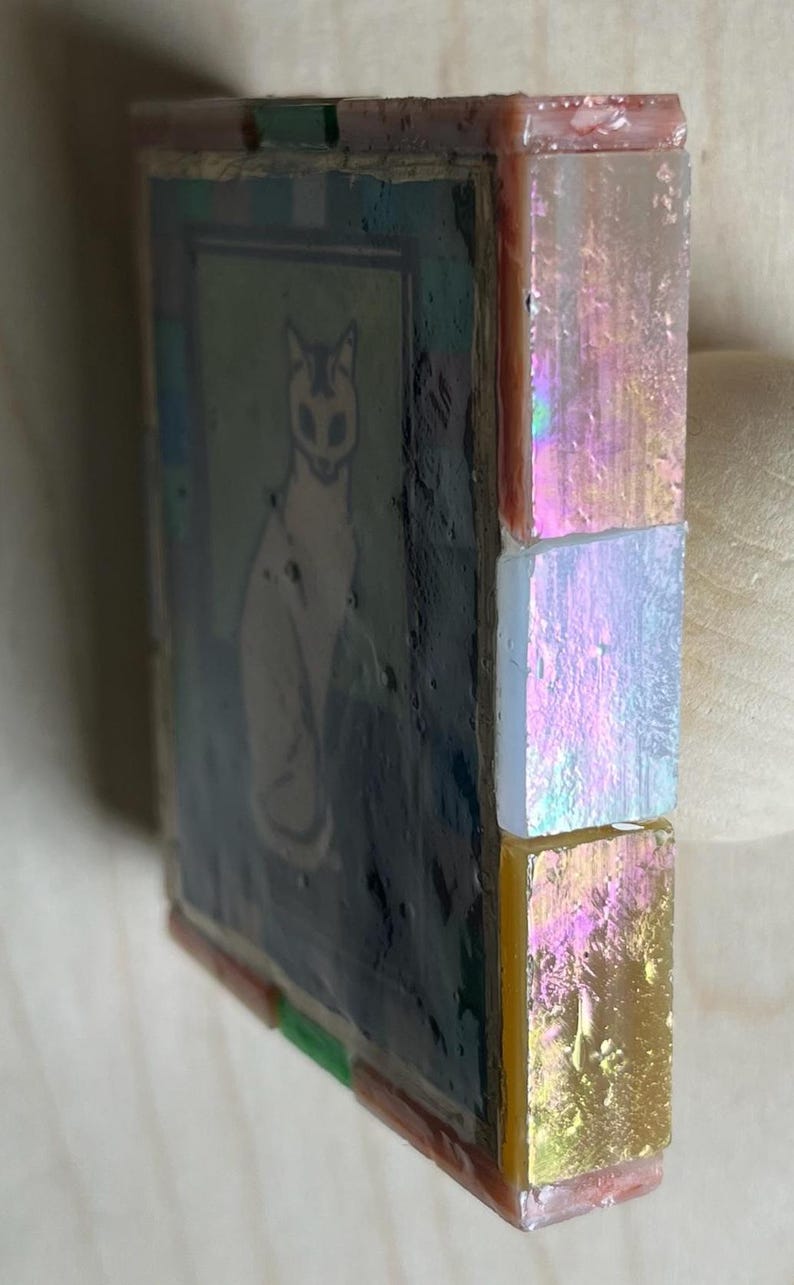 May include: A square mosaic tile featuring a central illustration of a cat on a dark background. The side edge is decorated with three vertically stacked, iridescent, rectangular glass tiles in shades of pink, white, and yellow-gold.