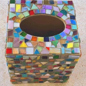 May include: A square tissue box cover decorated with a mosaic of multicolored glass tiles in shades of red, blue, green, yellow, and purple. The tiles are set in light gray grout, and the top features a large, centered oval opening for tissues.