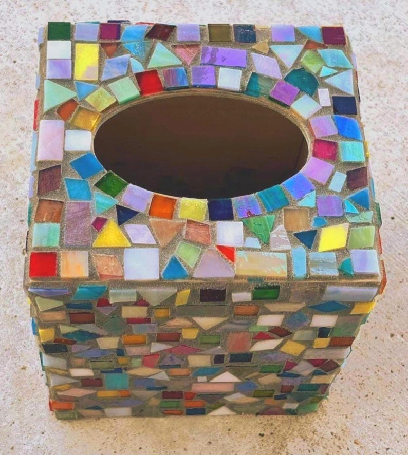 May include: A square tissue box cover decorated with a mosaic of multicolored glass tiles in shades of red, blue, green, yellow, and purple. The tiles are set in light gray grout, and the top features a large, centered oval opening for tissues.