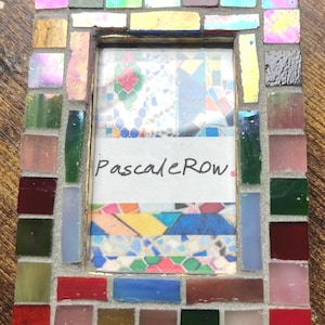 May include: A rectangular picture frame decorated with a mosaic of small, multi-colored glass tiles in shades of red, blue, green, pink, and gold. The center of the frame contains a white insert with the handwritten text PascaleRow in black ink.