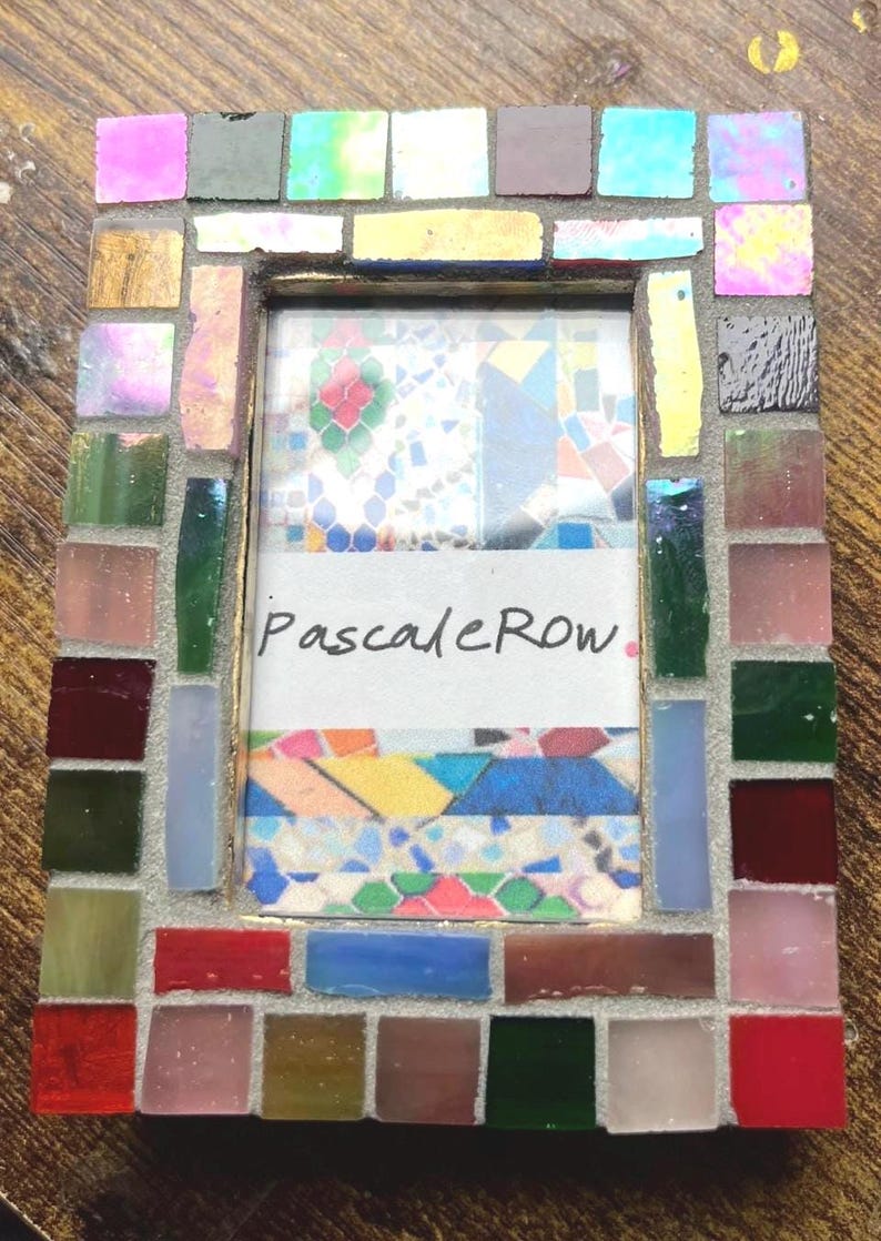May include: A rectangular picture frame decorated with a mosaic of small, multi-colored glass tiles in shades of red, blue, green, pink, and gold. The center of the frame contains a white insert with the handwritten text PascaleRow in black ink.