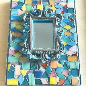 May include: A rectangular wall mirror featuring a central ornate blue and gold frame surrounded by a mosaic border of multi-colored glass tiles in shades of blue, green, yellow, and pink, set against a light gray grout background.