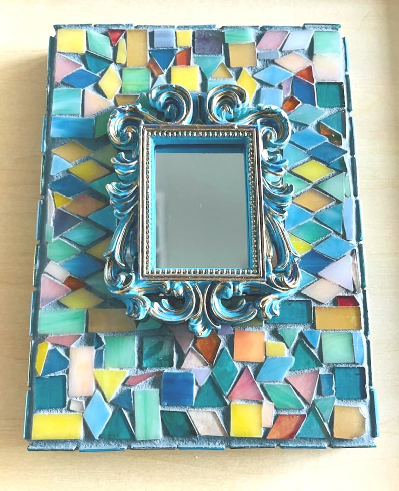 May include: A rectangular wall mirror featuring a central ornate blue and gold frame surrounded by a mosaic border of multi-colored glass tiles in shades of blue, green, yellow, and pink, set against a light gray grout background.