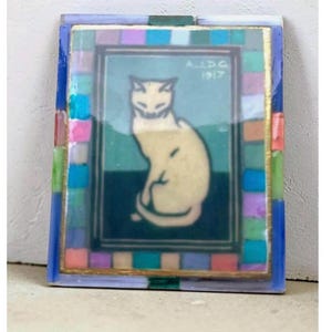 May include: A rectangular decorative tile featuring a central illustration of a seated cream-colored cat against a teal and dark green background. The text A.J.D.G. 1917 is printed in the upper right corner. A border of small, multicolored glass tiles surrounds the image.