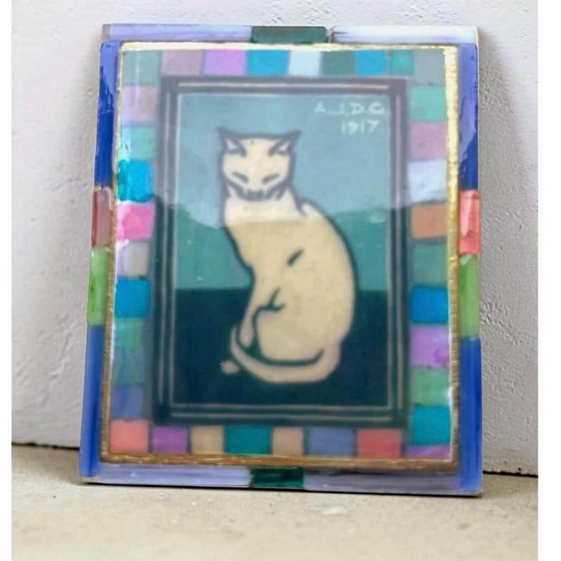 May include: A rectangular decorative tile featuring a central illustration of a seated cream-colored cat against a teal and dark green background. The text A.J.D.G. 1917 is printed in the upper right corner. A border of small, multicolored glass tiles surrounds the image.