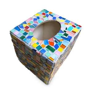 May include: A square tissue box cover decorated with a colorful mosaic pattern of small, irregular glass tiles in shades of blue, green, yellow, orange, pink, and white, featuring a large oval opening on the top surface for dispensing tissues.
