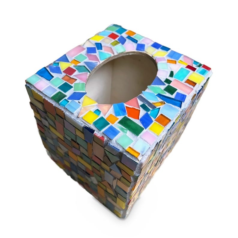 May include: A square tissue box cover decorated with a colorful mosaic pattern of small, irregular glass tiles in shades of blue, green, yellow, orange, pink, and white, featuring a large oval opening on the top surface for dispensing tissues.