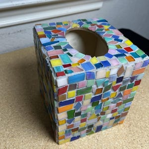 May include: A square tissue box cover decorated with a colorful mosaic pattern made of small, multi-shaped glass tiles in shades of red, orange, yellow, green, blue, and pink, set in light gray grout with a circular opening on the top surface.