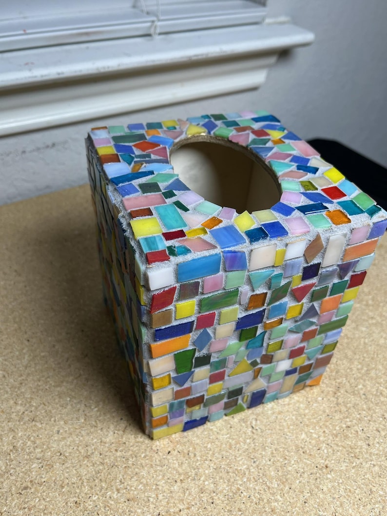 May include: A square tissue box cover decorated with a colorful mosaic pattern made of small, multi-shaped glass tiles in shades of red, orange, yellow, green, blue, and pink, set in light gray grout with a circular opening on the top surface.