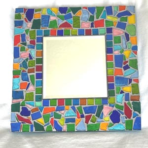 May include: A square wall mirror featuring a decorative frame composed of multi-colored glass mosaic tiles in shades of blue, green, red, orange, yellow, and pink, all set within a light gray grout, surrounding a central reflective glass pane.