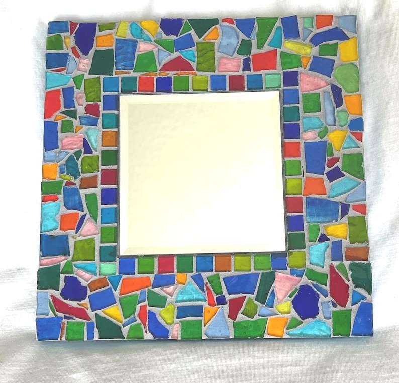 May include: A square wall mirror featuring a decorative frame composed of multi-colored glass mosaic tiles in shades of blue, green, red, orange, yellow, and pink, all set within a light gray grout, surrounding a central reflective glass pane.