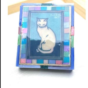 May include: A rectangular decorative box featuring a central illustration of a white cat on a blue background with the text A.J.D.G 1917. The border is composed of small, multicolored square tiles in shades of pink, blue, green, and purple.