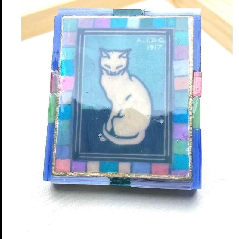 May include: A rectangular decorative box featuring a central illustration of a white cat on a blue background with the text A.J.D.G 1917. The border is composed of small, multicolored square tiles in shades of pink, blue, green, and purple.