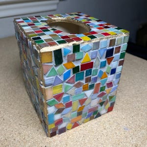 K&ouml;nnte beinhalten: Ein quadratischer Taschentuchbox-&Uuml;berzug, verziert mit einem bunten Mosaik aus kleinen Glasfliesen in verschiedenen Formen wie Dreiecken, Quadraten und Rechtecken. Die Kanten sind mit einer goldfarbenen Umrandung an der &Ouml;ffnung versehen.