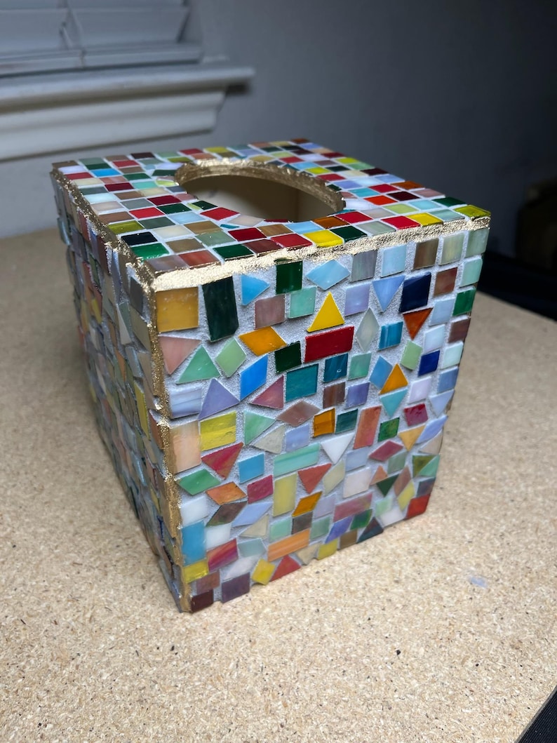 K&ouml;nnte beinhalten: Ein quadratischer Taschentuchbox-&Uuml;berzug, verziert mit einem bunten Mosaik aus kleinen Glasfliesen in verschiedenen Formen wie Dreiecken, Quadraten und Rechtecken. Die Kanten sind mit einer goldfarbenen Umrandung an der &Ouml;ffnung versehen.