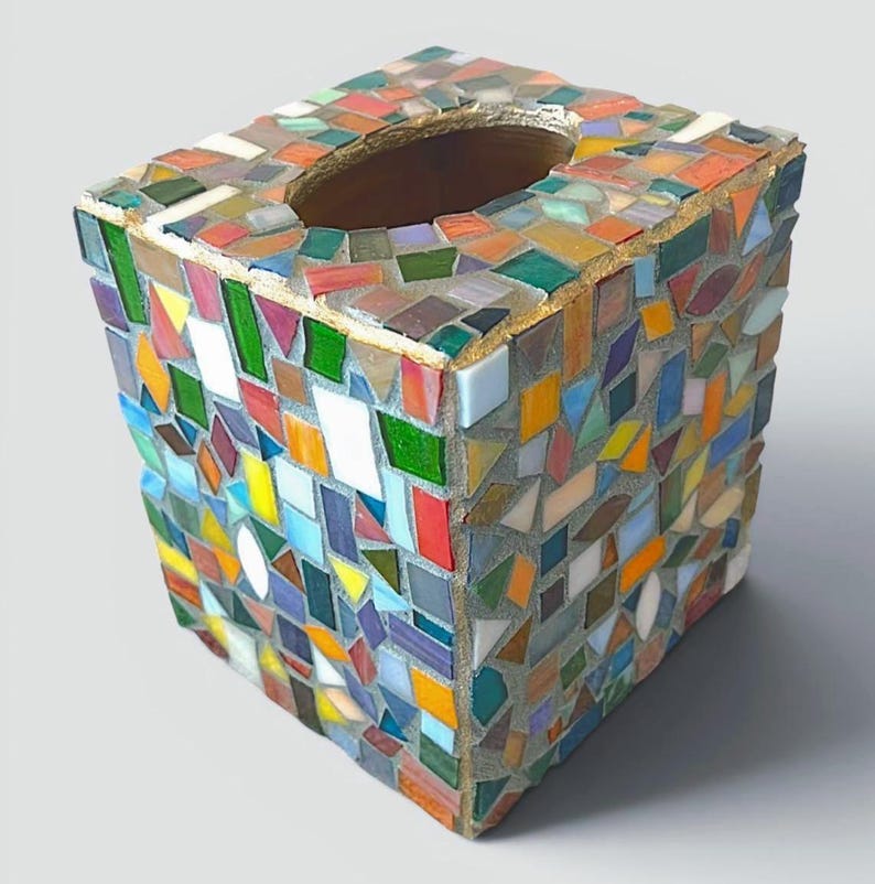 May include: A square tissue box cover featuring a multi-coloured mosaic design made of small, irregular glass tiles in shades of red, blue, green, yellow, and white, set into grey grout with a smooth, oval-shaped opening on the top surface.