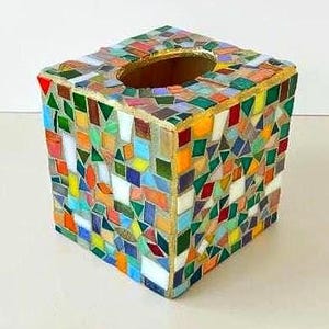 May include: A square tissue box cover featuring a multi-coloured mosaic pattern made of small, irregular glass tiles in shades of red, orange, yellow, green, blue, and white. The top surface has a central oval opening with a gold-toned border.