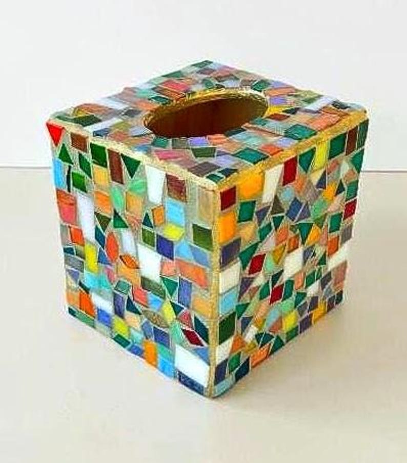May include: A square tissue box cover featuring a multi-coloured mosaic pattern made of small, irregular glass tiles in shades of red, orange, yellow, green, blue, and white. The top surface has a central oval opening with a gold-toned border.