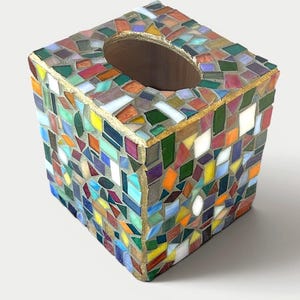 May include: A square tissue box cover featuring a colourful mosaic design made of various small, irregularly shaped glass pieces in shades of red, blue, green, yellow, and white, with gold-toned grout lines and a central oval opening.