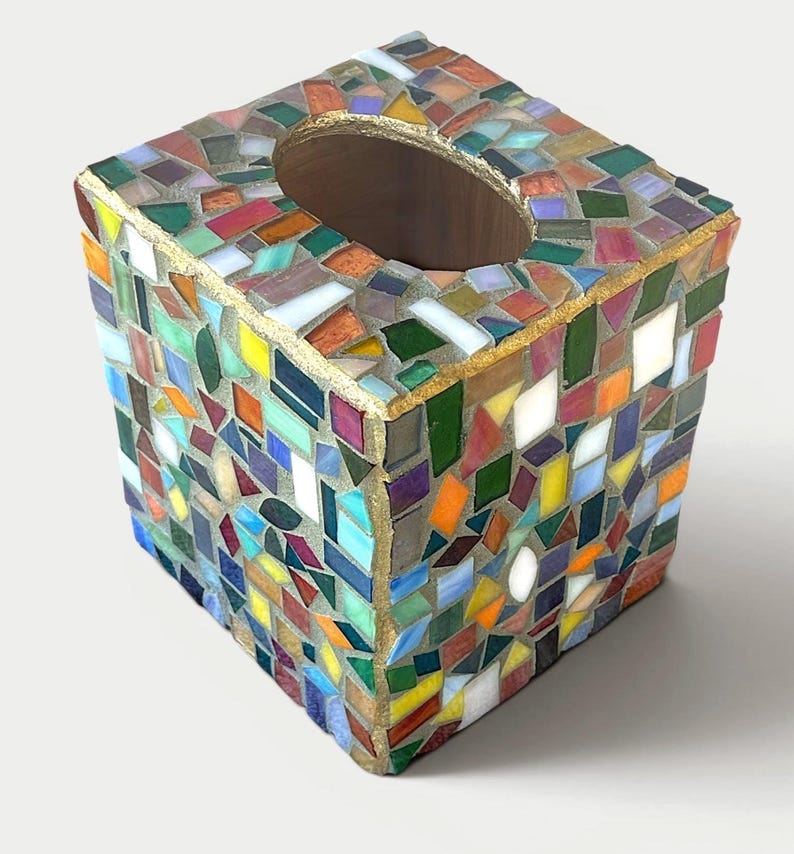May include: A square tissue box cover featuring a colourful mosaic design made of various small, irregularly shaped glass pieces in shades of red, blue, green, yellow, and white, with gold-toned grout lines and a central oval opening.