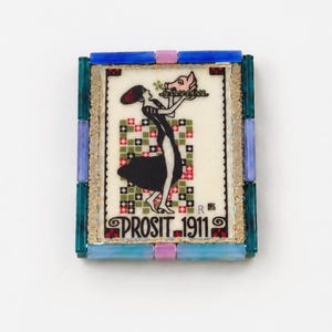 May include: A rectangular brooch featuring a central illustration of a figure in a black dress holding a tray with a pig head. The text PROSIT 1911 is printed at the bottom. The frame is composed of blue, teal, and lavender glass tiles.