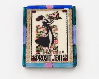 May include: A rectangular brooch featuring a central illustration of a figure in a black dress holding a tray with a pig head. The text PROSIT 1911 is printed at the bottom. The frame is composed of blue, teal, and lavender glass tiles.