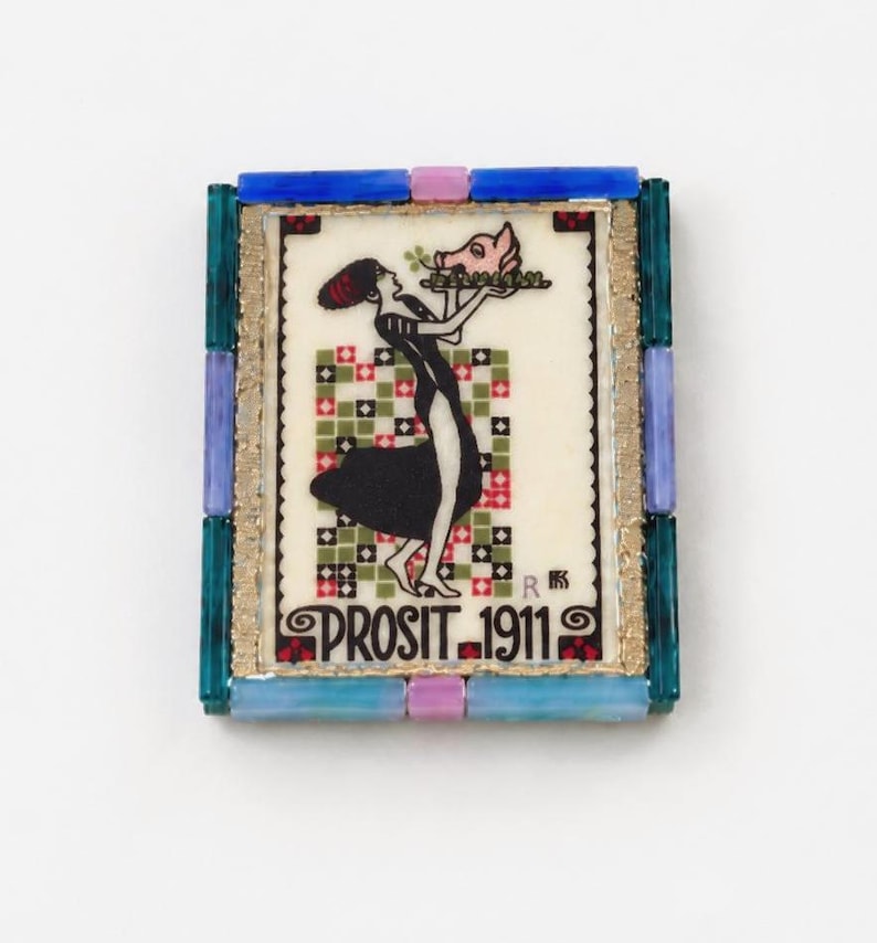 May include: A rectangular brooch featuring a central illustration of a figure in a black dress holding a tray with a pig head. The text PROSIT 1911 is printed at the bottom. The frame is composed of blue, teal, and lavender glass tiles.