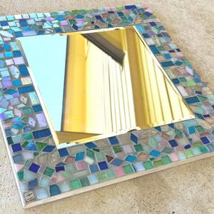 Op de afbeelding: Een vierkante spiegel met een decoratieve rand bestaande uit kleine, veelkleurige glazen moza&iuml;ektegels in tinten blauw, groen, roze en iriserend wit. Het centrale reflecterende oppervlak is glad en helder, omlijst door het getextureerde moza&iuml;ek.