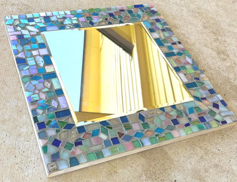 Op de afbeelding: Een vierkante spiegel met een decoratieve rand bestaande uit kleine, veelkleurige glazen moza&iuml;ektegels in tinten blauw, groen, roze en iriserend wit. Het centrale reflecterende oppervlak is glad en helder, omlijst door het getextureerde moza&iuml;ek.
