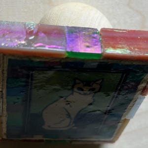 May include: A square decorative stamp featuring a central image of a cat on a dark background, framed by iridescent, multi-colored glass tiles in shades of pink, green, and purple, attached to a smooth, light-colored wooden handle base.