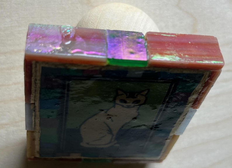 May include: A square decorative stamp featuring a central image of a cat on a dark background, framed by iridescent, multi-colored glass tiles in shades of pink, green, and purple, attached to a smooth, light-colored wooden handle base.
