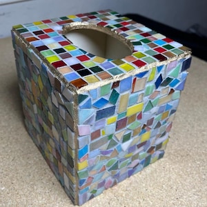 K&ouml;nnte beinhalten: Ein quadratischer Taschentuchbox-&Uuml;berzug, verziert mit einem bunten Mosaik aus kleinen Glasfliesen in Rot, Blau, Gelb, Gr&uuml;n und Wei&szlig;. Die Kanten und die ovale &Ouml;ffnung an der Oberseite sind mit einer strukturierten, goldfarbenen Metallkante versehen.