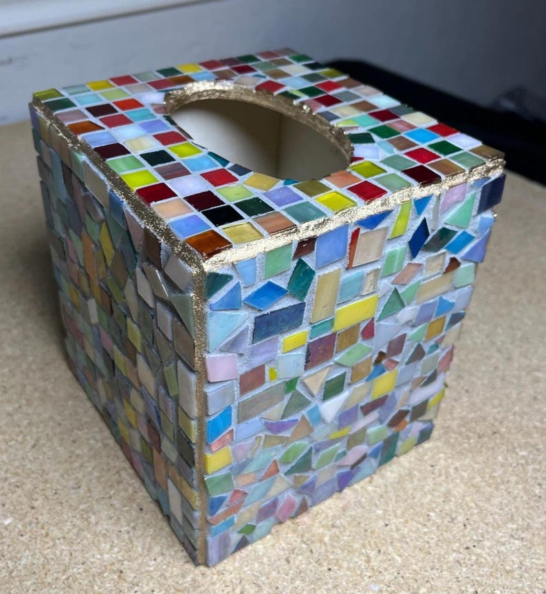 K&ouml;nnte beinhalten: Ein quadratischer Taschentuchbox-&Uuml;berzug, verziert mit einem bunten Mosaik aus kleinen Glasfliesen in Rot, Blau, Gelb, Gr&uuml;n und Wei&szlig;. Die Kanten und die ovale &Ouml;ffnung an der Oberseite sind mit einer strukturierten, goldfarbenen Metallkante versehen.