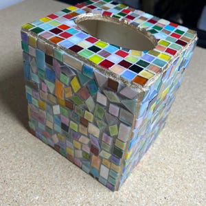 K&ouml;nnte beinhalten: Ein quadratischer Taschentuchbox-Bezug, verziert mit einem bunten Mosaikmuster aus kleinen Glasfliesen in Rot, Gelb, Blau, Gr&uuml;n und Wei&szlig;. Die Oberseite hat eine ovale &Ouml;ffnung mit goldfarbenem Rand, und die Seiten sind mit unregelm&auml;&szlig;igen geometrischen Fliesenst&uuml;cken bedeckt, die in Fugenm&ouml;rtel eingebettet sind.