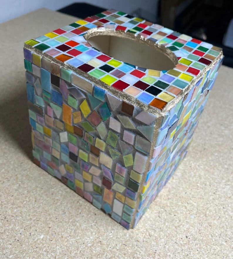 K&ouml;nnte beinhalten: Ein quadratischer Taschentuchbox-Bezug, verziert mit einem bunten Mosaikmuster aus kleinen Glasfliesen in Rot, Gelb, Blau, Gr&uuml;n und Wei&szlig;. Die Oberseite hat eine ovale &Ouml;ffnung mit goldfarbenem Rand, und die Seiten sind mit unregelm&auml;&szlig;igen geometrischen Fliesenst&uuml;cken bedeckt, die in Fugenm&ouml;rtel eingebettet sind.