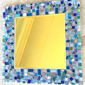 Op de afbeelding: Een vierkante spiegel met een decoratieve lijst bestaande uit veelkleurige glazen moza&iuml;ektegels in tinten blauw, groen, wit en zachtroze. Het centrale reflecterende oppervlak is glad en goudkleurig, omringd door een rand van diverse geometrische vormen.